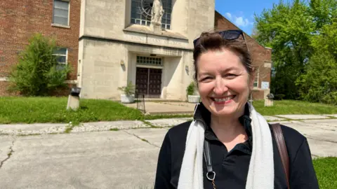 Mary Simons smiling outside of a stone fronted church with weeds in the background.