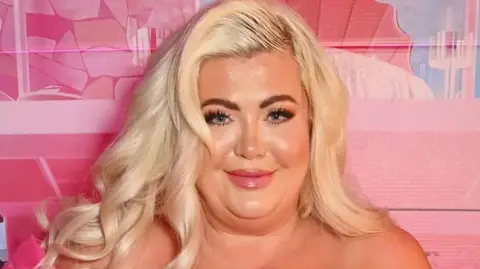 Gemma Collins is photographed looking into the camera at an event. She has dark brown eyes, long eyelashes and long, bleached blonde hair. The background wall behind her is different shades of pink.