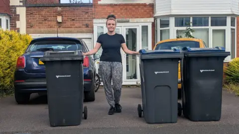 Tonia Dunn A woman with dark hair tied back, wearing a black T-shirt, trainers and patterned trousers stands next to three black wheelie bins. A house and two cars are behind her on a driveway.