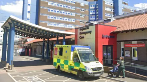 The exterior of the University Hospital of North Tees. The brick building has signs on it that read: "Urgent & Emergency Care". There is an ambulance parked out front and a paramedic pushing a wheelchair.