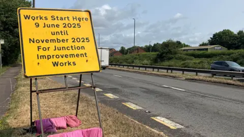 A yellow roadworks sign with 'work starts here 9 June 2025 until November 2025 for junction improvement works'. 
The sign is being held in position by a number of pink sand bags at the base. The sign is on the side of a dual carriageway. 