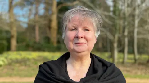 Ann Cavozzi is wearing a black top and has grey hair. In the background a wooded area is visible.