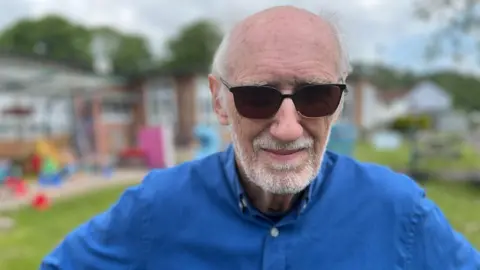 A man looking at the camera. He is wearing a blue shirt and has a grey beard and moustache, it is stubbly. He has dark shades on and behind him is the school, which is slightly out of focus.