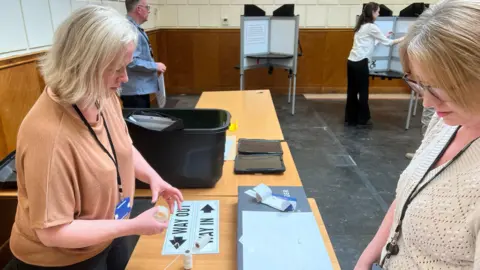 Mousumi Bakshi/BBC Three people are setting out tables, boxes and polling booths.