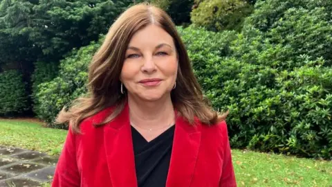 McLaughlin is smiling at the camera. She is wearing a red blazer and black top. she has shoulder length brown hair.