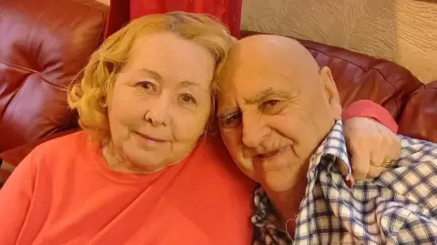 Tracy Faram Shipton Elizabeth and her husband John are photographed sat on a red sofa from the shoulders up. Elizabeth has her arm around his shoulder. 