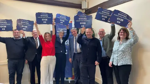 A group of people are smiling into the camera and holding up blue pieces of paper which say Grimsby GY28 on them. They are wearing work attire and standing in a room in front of a poster behind them.