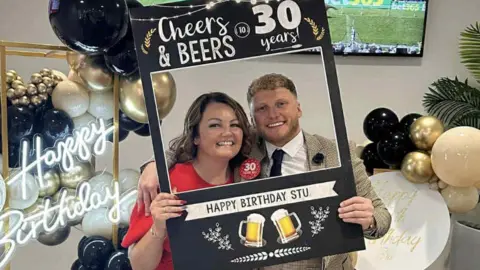 Debra Wright (left) with long dark brown hair wearing a red dress poses in a gap in a 30th cardboard birthday card designed for selfies along with her son Stu McDonald who has light brown hair and stubble wearing a suit. They are both smiling. 