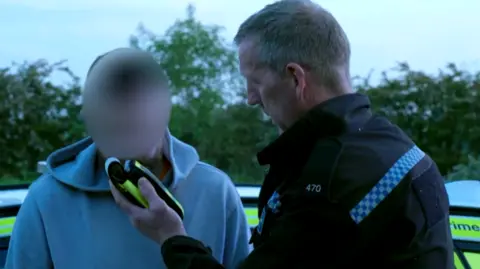 A still image taken from a video shows a Northamptonshire Police officer carrying out a roadside breathe test on a motorist in a grey hoodie.