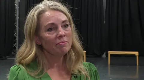 A woman in a green dress with long blonde hair in a theatre with a black backdrop.