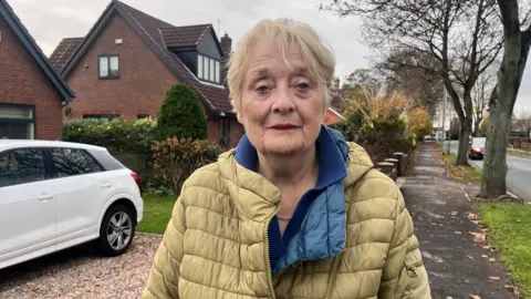 Mary stands outside her home in Cottingham wearing a yellow jacket and blue top and stares directly at the camera, looking concerned. Behind her is a gravel drive and white car.