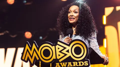 Kanya King speaking on stage at a lectern with a large yellow Mobo Awards logo at the 2025 ceremony
