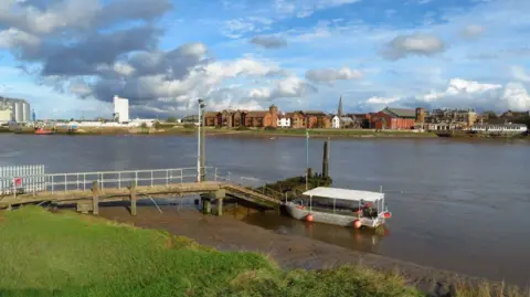 A small ferry boat is parked at a river jetty with the grassbank in the foreground and buildings lining the other side of the river in the distance