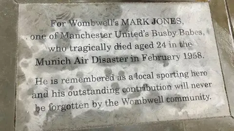 Victoria Scheer/BBC The image shows a stone memorial plaque set into the ground. The plaque is rectangular and bordered by smooth stone tiles. The inscription pays tribute to Mark Jones, noting his connection to Wombwell and his role as one of Manchester United’s Busby Babes. It explains that he died at the age of 24 in the Munich Air Disaster in February 1958. The plaque also states that he is remembered as a local sporting hero and that his contribution will always be valued by the Wombwell community.