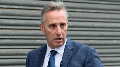 Ian Paisley - a man with short grey hair wearing a striped blue suit, a blue tie and a white collared shirt is standing in front of grey metal shutters.