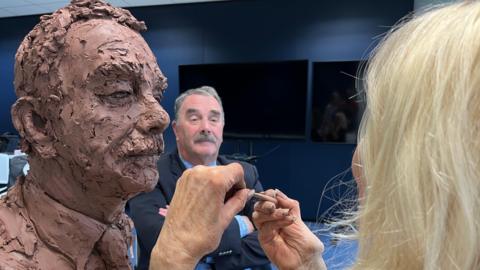 A clay sculpture of a man, who is sat in the middle further back in soft focus. Two hands appear to be working on the sculpture.