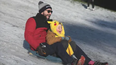 Pacemaker A man in a red and black puffer jacket, a grey bobble hat and a pair of black sunglasses is riding on a sleigh with a young child dressed in a Pikachu Pokémon costume. Both have their mouths open as they sled down the snow covered hill.