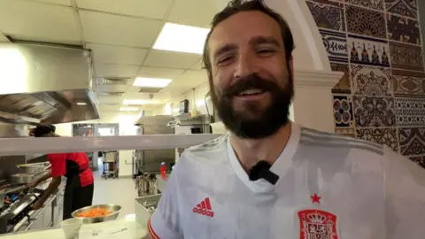 Sarah-May Buccieri / BBC Amador Abruneiras Jr., el gerente del restaurante, sonríe y viste un uniforme blanco de fútbol de España. Está de pie frente a la cocina blanca y luminosa del restaurante. Al fondo, un trabajador de cocina cocina en muebles cromados.