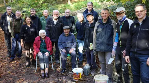 A group of volunteers standing by a wall in a forest estate, two are sitting down the rest of standing up, they are all looking at the camera and smiling. They are in a forest area, with brown leaves on the ground foliage behind them. 