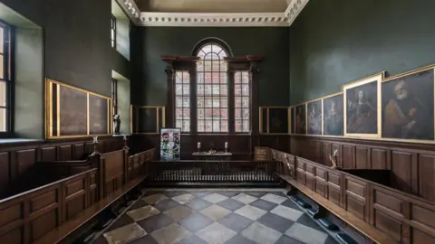 A small chapel with a large arched window at the front and three smaller windows to the left-hand side. The windows are made of smaller oblong-shaped glass panels. There is wooden panelling around the bottom third of the walls, and wooden enclosed pews either side of a white and black stone floor. There is a small altar front & centre with two candles and a cross on it. There are oil paintings around the chapel above the wood panelling.