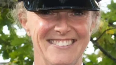 North Yorkshire Police A police officer in her forties, smiling directly at the camera. She is wearing a police cap which comes down to just above her eyes. She is pictured against the backdrop of some trees.