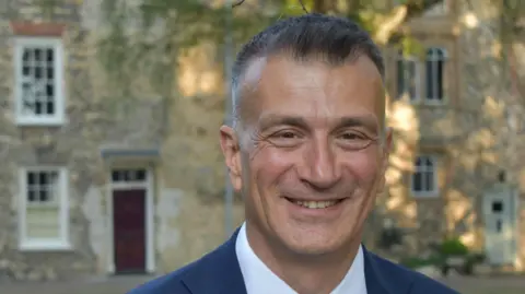 Dr Nikos Savvas smiles at the camera as he stands outside in front of several school buildings. He has short black and grey hair and wears a blue suit with a white shirt underneath. 