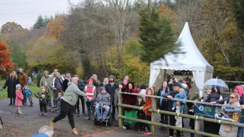 Catsfield Christmas Tree Farm Roughly two dozen people stood behind a wooden fence next to a road. They watch as a man throws a Christmas tree up the road. Behind them is a white gazebo and woodland.