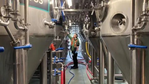 Alex Pope/BBC A man standing between large metal containers in a brewery. He is dressed in black trousers and green top with a high-vis jacket and ear defenders on. He is holding one arm up. There are pipes around him. 