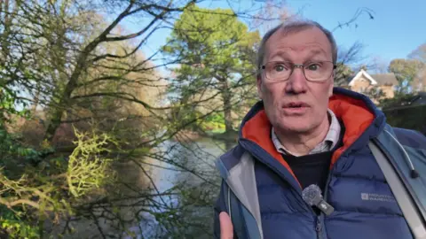 Prof John Sear in a blue anorak standing alongside a river with trees on either side