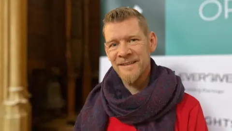 Shariqua Ahmed/BBC Henning Grosse Ruse-Khan wearing a red shirt and a purple scarf, smiling for camera.