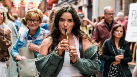 A woman with long brown hair, playing a tin whistle. There is a crowd of people behind her. 