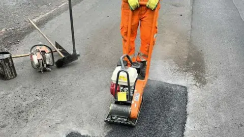 North Lincolnshire Council A worker in orange overalls pushes a pothole-repairing machine, which resembles a lawnmower, over a patch of newly laid black asphalt on a grey road. Brushes, a bucket and more machinery can be seen.