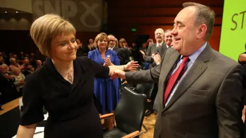 PA Media A man and woman have arms outstretched as they prepare to hug on a stage with clapping people behind them. The woman, with short fair hair, is wearing a black dress. The man, with short grey hair, is wearing a grey suit and red tie. 