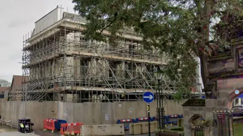 Google The hotel that is surrounded by a fence and covered in scaffolding. A tree can be seen hanging overhead and there is also a road sign in sight. 