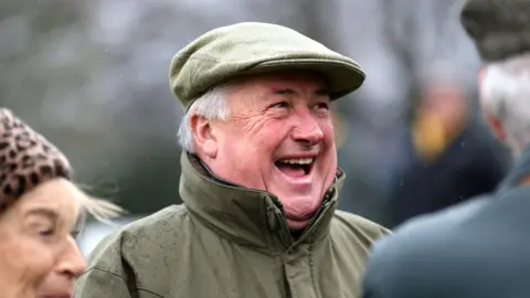 PA Media Paul Nicholls smiling with his mouth open, wearing a khaki coloured coat and cap