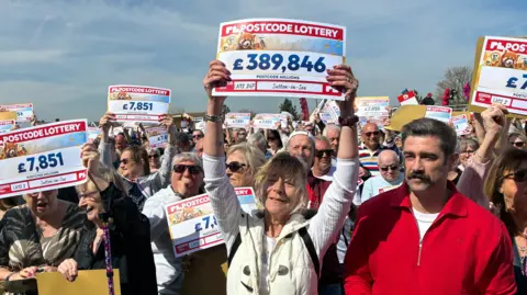 A large crowd of people stand holding replica cheques above their heads, with a woman with blonde hair in the foreground holding a sign stating £389,846.