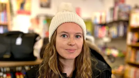 Martin Giles/BBC A woman stares at the camera, she wears a white hat, black coat and black and white stripey top. In the background are the rails and shelves of a charity shop. 