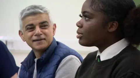 Sadiq Khan is seen sitting next to a PRU pupil. He is smiling as she appears to be speaking to a wider group not in view.