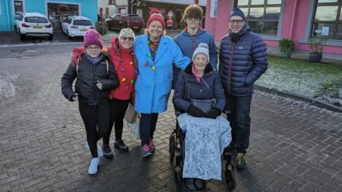 Denise Baxter A family smile for the camera while walking down a street. The group consists of three women, including one sitting in a wheelchair, a man with glasses and a hat, a male teenager and a young girl.