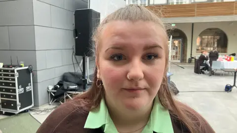 A teenage girl, wearing a green shirt and a brown jumper. She has long brown hair, tied back and has earrings and a nose ring. 
