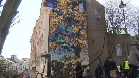 A large mural of a painted tree on the side of a building in Newton Abbot. The tree has yellow and gold leaves coming off the branches, and the background has light and dark tones of blue. There are people stood around it, including people wearing yellow hi-vis.