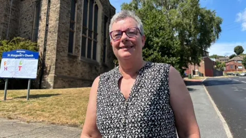 Simon Thake A middle aged woman with short grey hair stands on the pavement outside a church