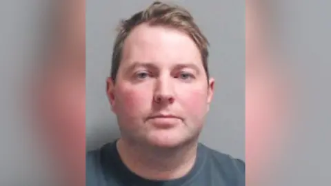 PSNI Jonathan Creswell has short brown hair parted to the side and light blue eyes. He is wearing a blue/grey t-shirt and is staring straight ahead in a police mugshot. 