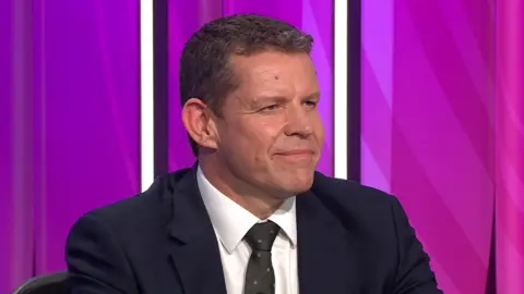 A head and shoulders photograph of Rhun ap Iorwerth sitting, smiling, on the Question Time set with a purple background behind him and wearing a dark suit and tie and a white shirt.