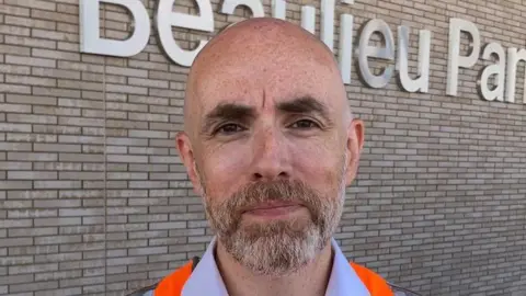 Joshua Holmes-Bright/BBC Martin Beable wearing an orange hi-vis vest over a salmon pink shirt. He is smiling in front of a brick wall that has silver lettering on it that reads "Beaulieu Park".