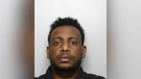 A police mugshot of Daniel Hayelom, a man with black afro hair and stubble. He is staring into the camera.