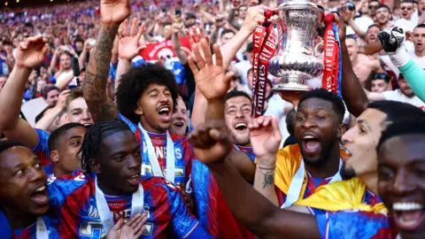 Crystal Palace FC players gather together on the pitch in front of crowds, all smiling, one holding the FA Cup trophy in the air 