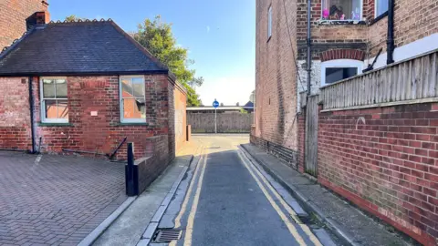 A narrow single-track road, with double-yellow lines and narrow pavements, passes between a one-storey, red-brick building and a three-storey red-brick house.