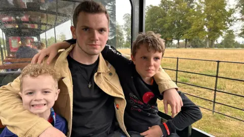 Family handout Kacey and his older brothers Morgan and Brody are pictured sitting in the carriage of a novelty train with their arms around each other.  There is a field and trees in the background. Kacey is smiling and the other two are looking quite serious.