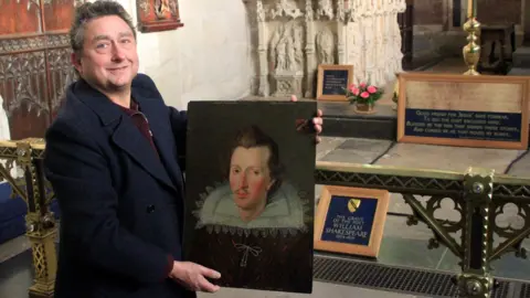 Madoc Roberts Steven holds the painting up for view in a church that has a plaque saying "he Grave of the Poet William Shakespeare". There are large flagstones on the floor and a brass ornate rail. Steven wears a blue woollen coat and maroon shirt